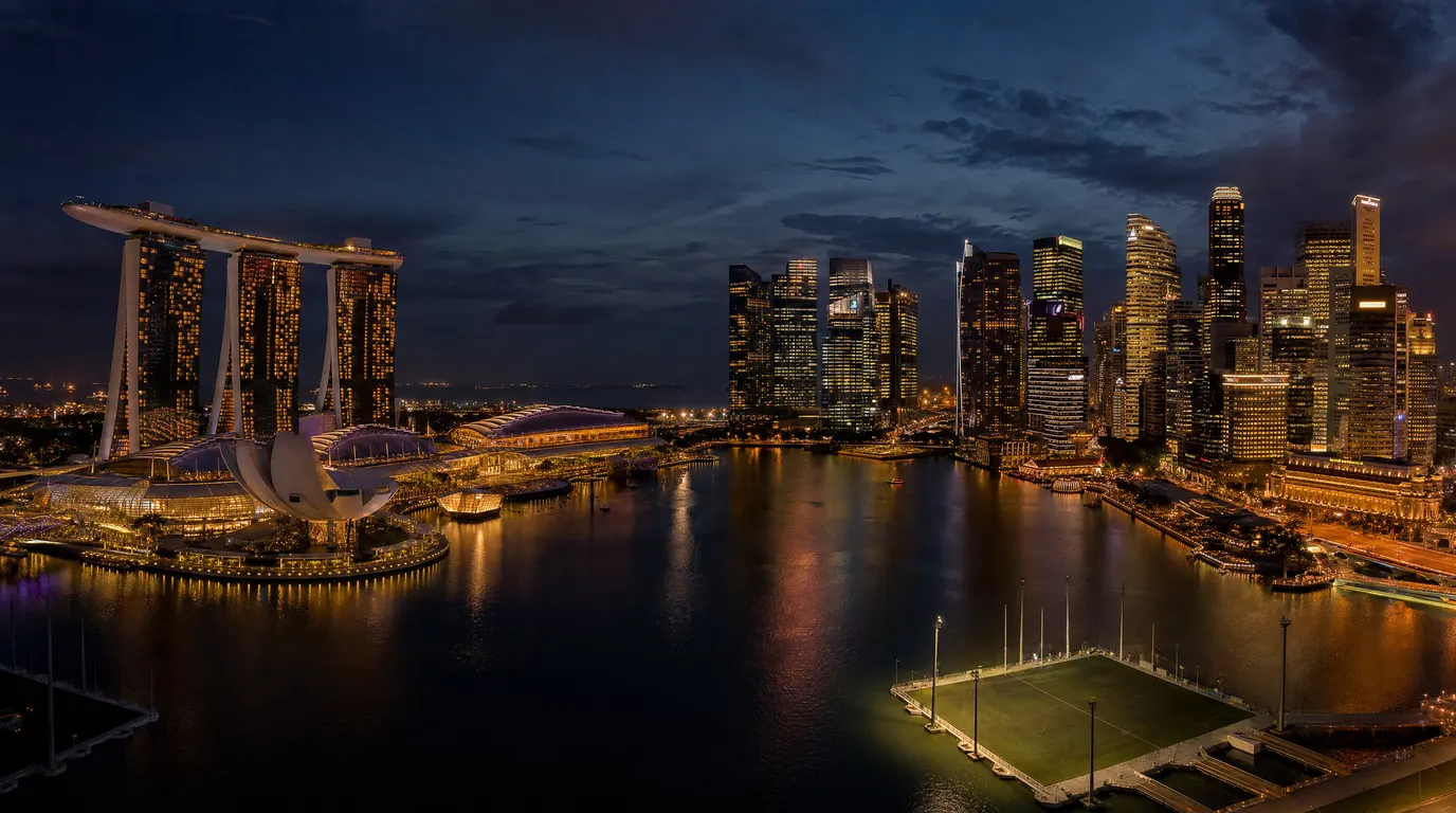 Aerial cinematic view of Singapore's Marina Bay financial district at dusk, warm amber tower lights reflected on calm water, dark navy sky, long lens compression, no text or signage